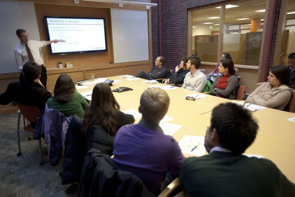 Students in a classroom around a long table watching an instructor pointing to his screen.